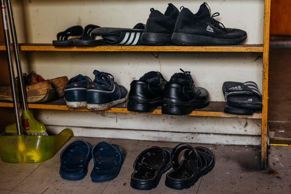 The shoes of multiple boarding house occupants. Tenants hang clothes above their beds.
